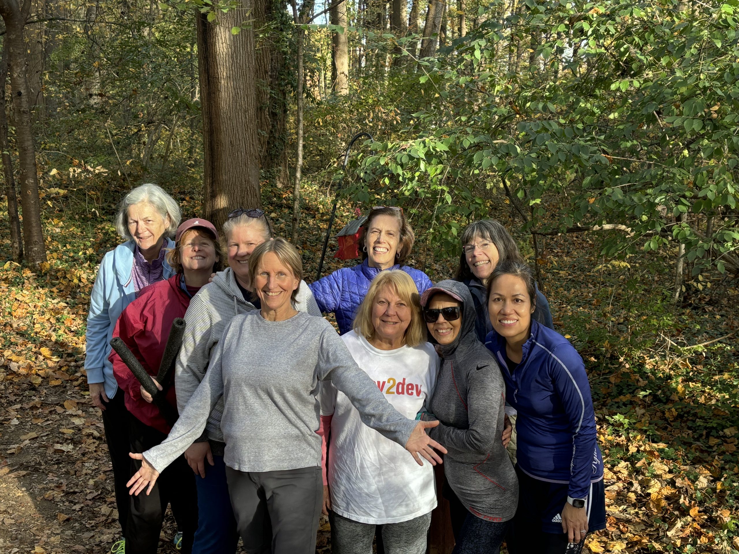 A group of nine middle-aged women looking happy in the woods after a workout. Women over 50. Health coach.